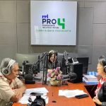 Three radio hosts wearing headphones sit around a wooden table with multiple microphones in a studio with a logo wall behind them